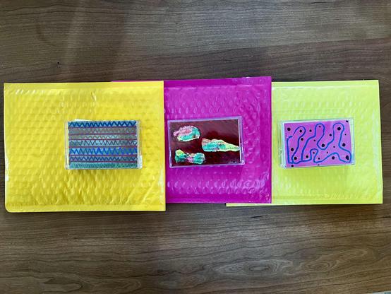 These are hand-decorated cassette tapes displayed on colorful bubble pack mailers.

The three cassette tapes are positioned on vibrant padded envelopes:

Left (Yellow mailer): A cassette tape decorated with a geometric pattern featuring blue and turquoise triangular designs in rows.

Middle (Magenta/hot pink mailer): A cassette tape with an abstract collage-style decoration in dark red/maroon with bright turquoise and pink accents, and some text elements.

Right (Lime green mailer): A cassette …