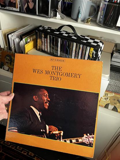 This photo shows a classic jazz vinyl album being held up in a personal music collection space.

The Album:
"The Wes Montgomery Trio" on the Riverside Records label

Features the legendary jazz guitarist Wes Montgomery

The cover shows Montgomery in formal attire (dark suit and tie) playing his guitar

Distinctive orange/golden yellow border framing the album

Classic jazz photography style with dramatic lighting highlighting Montgomery's profile and instrument

The Setting:
The album is being …