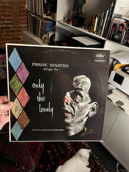 This photo shows a classic Frank Sinatra vinyl album being held up in a music collection space.

The Album:
"Frank Sinatra Sings for Only the Lonely"

Released on Capitol Records (logo visible in top right)

Orchestra conducted by Nelson Riddle

One of Sinatra's most celebrated concept albums from 1958

Album Design:
Striking black cover with minimalist, elegant design

Dramatic high-contrast portrait of Sinatra's profile looking upward

The title "only the lonely" written in elegant white scri…
