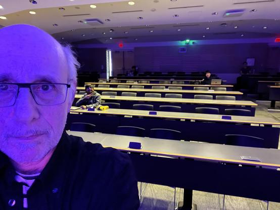 A selfie taken from the front of a large lecture hall, mostly empty. The person in the foreground — wearing glasses and a dark top — is lit by a striking purple/blue light that gives the whole scene a moody, somewhat dramatic atmosphere.

In the background, the tiered rows of gray chairs and long desks stretch back through the hall, with only a handful of students present — one nearby giving a peace sign, and a couple more scattered toward the back, one with a laptop open.

The vibe is somewher…
