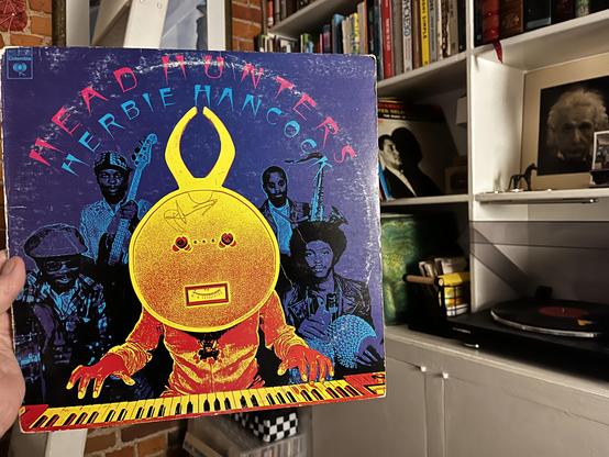 This photo shows a striking album cover with bold psychedelic artwork from jazz legend Herbie Hancock.
The Album:
"Head Hunters" by Herbie Hancock
Released on Columbia Records (1973)
One of the best-selling jazz albums of all time
Album Design:
Vivid purple/deep blue background
Title in pink and cyan lettering arcing across the top
Central figure: a stylized African-inspired mask/character in bright yellow, orange, and red gradient
The character has geometric features, circular head desiβ¦
