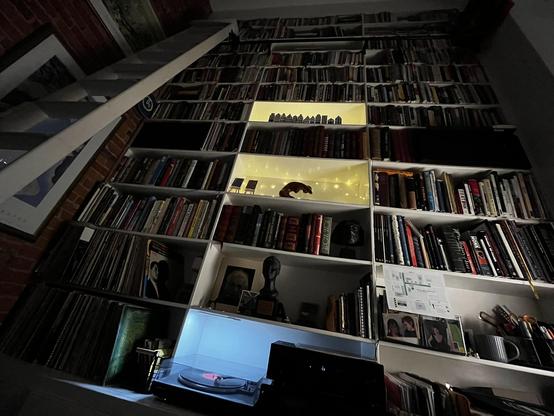 This dramatic photo captures an impressive floor-to-ceiling media library photographed from below, looking up at night.

The Collection:
Massive shelving system spanning the entire wall, packed with:

Hundreds of vinyl records organized vertically

Extensive book collection (hardcovers and paperbacks)

CDs and other media

Multiple sections reaching from floor to ceiling

Lighting & Atmosphere:
Dramatic string lights creating a warm yellow glow on two shelves

The lights highlight a city skylin…