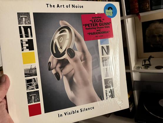 This photo shows a groundbreaking experimental music album with striking modernist artwork.

The Album:
"The Art of Noise - In Visible Silence"

Released on ZTT Records (1986)

Includes hit tracks: "Legs," "Peter Gunn" (featuring Duane Eddy), and "Paranoimia"

Album Design:
Iconic sculptural profile of a head in grayscale with stylized @ symbol embedded in it

Clean white border with minimalist design elements

Left side: vertical strip with black and white photographic images, yellow and red c…