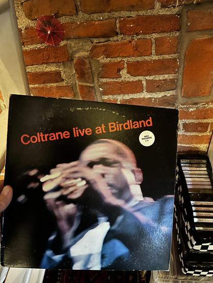 This photo shows one of the most legendary live jazz albums ever recorded.

The Album:
"Coltrane live at Birdland" by John Coltrane

Released on MCA Records (reissue of the original 1964 Impulse! recording)

Bold orange text against black background

Album Design:
Powerful live performance photograph of Coltrane

Captured mid-performance with intense expression

His hand near his face, deeply immersed in the music

Blue tones in his clothing

Motion blur suggesting the energy and movement of li…