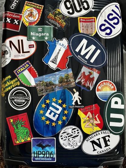 These photos show a well-traveled black hardshell suitcase covered with travel stickers and decals.

Michigan & Great Lakes:

MI (Michigan oval)
UP (Upper Peninsula)
Michigan - The Great Lakes State (large blue silhouette)
NF - Niagara Falls, Canada
Niagara (highway sign style)
Thunder Bay National Marine Sanctuary
Northern Michigan University Wildcats
HP906 (Upper Peninsula area code with bridge silhouette)

European Destinations:

Amsterdam (multiple - including postcard-style cityscape and m…