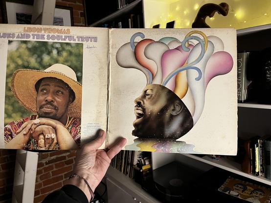 This photo shows a classic soul and R&B albums opened in a gatefold display, showcasing contrasting but equally striking cover art.

Left side (Album back side):
"Leon Thomas - Blues and the Soulful Truth"

Portrait of Leon Thomas wearing a wide-brimmed straw hat

Colorful patterned shirt

Contemplative pose with hands clasped

Natural outdoor setting with green bokeh background

Warm, earthy tones

Right side (Album front side):
Beautiful psychedelic/spiritual artwork

Profile of a face tilted…