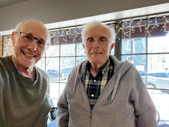 This warm photo shows two men sharing a friendly moment in what appears to be a casual restaurant or café setting.

The People:
Left: A man [it's-a-me!] with a gray goatee wearing black-framed glasses and a gray-green crewneck sweater, smiling warmly at the camera

Right: An older gentleman with white hair wearing wire-rimmed glasses, a gray zip-up hoodie over a plaid flannel shirt, also smiling

The Setting:
Large windows with gridded panes letting in natural light

String lights with warm whi…