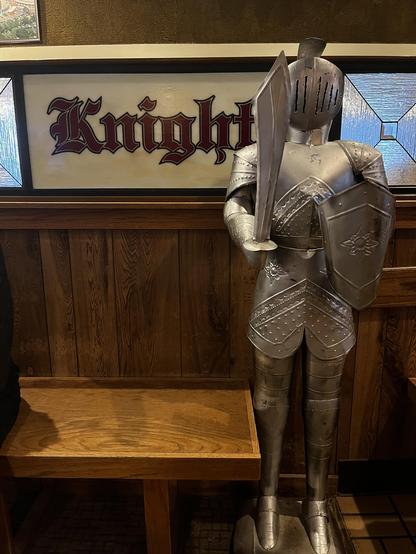 This photo shows medieval-themed restaurant decor.

The Knight:
A life-sized suit of armor standing guard with full plate armor, closed helmet, large shield with star emblem, sword at ready, and authentic metallic weathering.

The Signage:
Behind the knight: “Knight” in Gothic blackletter, burgundy text on cream background, flanked by blue stained glass-style panels.

The Setting:
Dark wood paneling, wooden bench seating, tile floor, olive/gold upper walls - classic pub atmosphere with medieval…