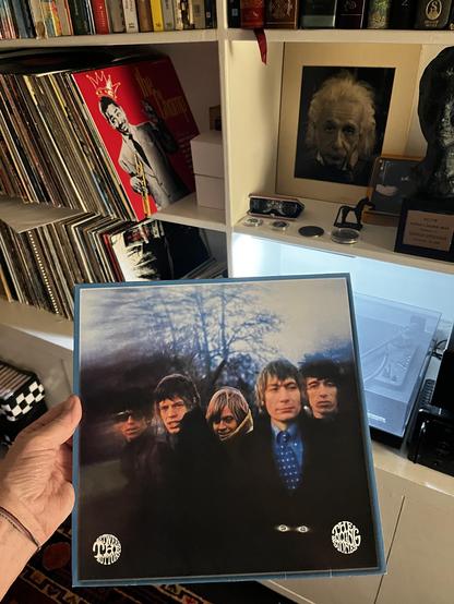 This photo shows "Between the Buttons," a classic Rolling Stones album.

The Album:
Iconic moody photograph of the young band

Four members posed outdoors at dusk with bare tree silhouettes

Dark clothing creating shadowy, atmospheric aesthetic

White Rolling Stones logo circles at bottom corners

The Setting:
White shelving system packed with records and memorabilia:

Extensive vinyl collection on the left

Bold red album cover visible (with crown imagery)

Einstein photograph in frame

Variou…