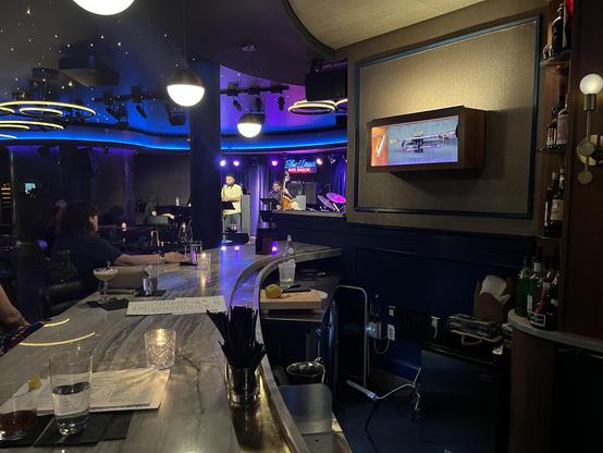 The Blue Llama looks terrific — that deep blue ambient lighting with the star-field ceiling gives it a real supper-club intimacy. You can see the neon “Blue Llama / Ann Arbor” sign glowing on the back wall of the stage, and on the right wall there’s a framed wooden case displaying a trumpet, a nice touch that sets the jazz-club atmosphere.

On stage, two musicians are visible: one standing at a music stand (a horn player at rest or between phrases), and another seated at the bass. The drum kit …