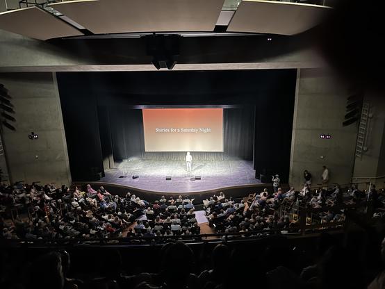 Wide view from the upper balcony of the Power Center for the Performing Arts in Ann Arbor, showing Ira Glass standing alone on stage under a spotlight, with a screen behind him reading 'Stories for a Saturday Night' on an orange-to-cream gradient. The seating bowl below is filled with audience members, and a photographer with a camera stands near the right edge of the stage.