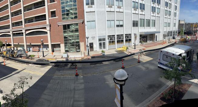 Elevated panoramic view of a downtown street under construction. A long black flexible conduit, roughly a block in length, snakes along the pavement, propped up on small wooden cradles and orange traffic cones. A John Deere backhoe sits at the curb near a brick parking structure on the left. The street has been cut open in patches, showing fresh asphalt repairs. Storefronts line the sidewalk, including one marked "BLOM," with bistro tables, bike racks, and string-light poles along the curb. A "…
