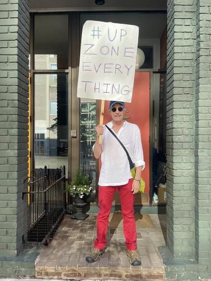 Man standing in a doorway with a red front door and green brick facade, holding a handmade cardboard sign on a wooden stick that reads "#UPZONE EVERYTHING" in purple marker. He wears a white linen shirt, red pants, sunglasses, and a navy baseball cap, with a crossbody bag over one shoulder. Potted flowers and a wrought iron railing sit beside the brick steps.
ALT by Claude