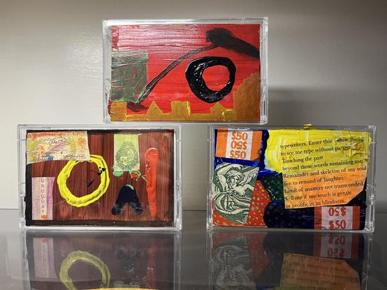 Three clear plastic cassette cases, each holding a small mixed-media artwork, stacked one on top and two below, against a plain wall, reflected in a dark glossy surface.

**Top:** A fiery red background with a thick, glossy black ring right of center. A long black brushstroke sweeps diagonally through it, and a cluster of dark strokes gathers in the upper right. A jagged ochre line along the bottom suggests a skyline, while a patch of muddy sage green breaks up the red on the left.

**Bottom le…