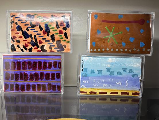 Four cassette cases in a 2x2 grid, painted on their inlay cards, against a plain wall, reflected in a dark glossy surface below.

Top left: Peach-orange background scattered with small painted marks in navy, black, silver, red, and green — teardrops, triangles, dashes, blobs — a lively confetti-like pattern with no single focal point.

Top right: Warm brown background with sky-blue teardrops around the border. A thick magenta line stretches across the top, ending in a rounded blob. A green X si…
