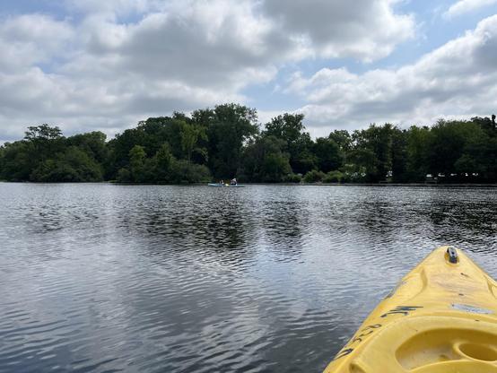 A view from the bow of a yellow kayak, looking across a calm, ripple-textured stretch of the Huron River. In the middle distance, a two-person kayak carries Lynn and grandson Max, paddles in motion, both wearing life vests and one in a wide-brimmed hat. A dense green tree line lines the far shore, with a hint of a parking area and dock visible through gaps in the foliage. Overhead, a mix of blue sky and heavy cumulus clouds fills most of the frame, their reflections breaking up on the water's s…