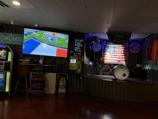 The inside of the Wagon Wheel bar in Ossineke, Michigan — a small-town tavern with a stage/live music setup. A big TV is showing France vs. England on Fox Sports (looks like a national anthem/pre-match moment, with both teams lined up on the pitch). There's a lit-up entertainment board listing trivia, karaoke, open mic nights, and band names for July/August.

To the right is a small stage with a Pearl bass drum, an American flag, and a "Wagon Wheel, Where Good Friends Come Around – Ossineke, Mi…
