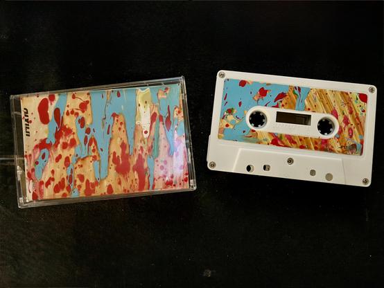 On the left, a clear plastic Norelco case, with a j-card slid in. On the right, the cassette itself lying on its side, white shell with matching artwork on the label.

The artwork is the same abstract paint-pour design on both the j-card and the cassette label — vertical drips and splatters in sky blue, tan/ochre, and red, with a few softer touches of pink, cream, and green. It has that marbled, poured-paint look, like the colors were layered and allowed to run vertically down the card before d…