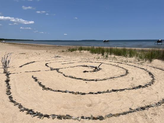 A stone spiral labyrinth on the sandy shore of Thunder Bay, laid out with flat dark shale pieces and a piece of driftwood marking the center. The view looks out across the bay toward Alpena, with calm blue water, a tree-lined shoreline in the distance, and a couple of small structures (what look like a swim platform and a dock) out on the water. Dune grass and a few bleached driftwood branches frame the scene in the foreground under a clear, mostly sunny sky.

ALT by Claude 