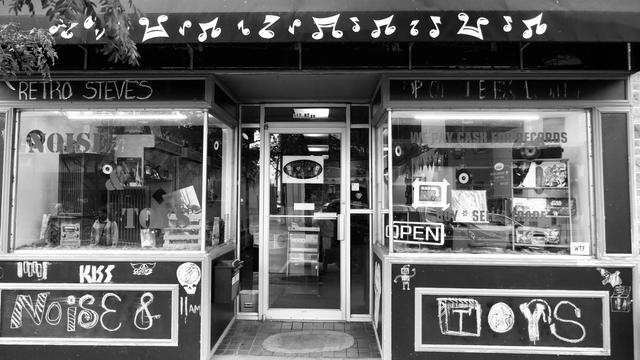 A storefront entrance is flanked by two big display windows painted with "NOISE & TOYS" and "WE PAY CASH FOR RECORDS," plus a hand-drawn "OPEN" sign and a Record Store Day Drops poster in the right window. The awning above is decorated with a row of oversized musical notes cut from what looks like white vinyl or paper, giving it a fun, handmade record-shop vibe. Chalkboard panels below each window are covered in band names and doodles — KISS, Aerosmith, a Grateful Dead skull-and-lightning-bolt,…