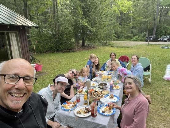 Aelfie with me in the foreground grinning stupidly at the camera.  Behind me stretches a full picnic table under the trees, mid-cookout (hot dogs, mac and cheese, chicken, ketchup and mustard bottles making the rounds).

Reading down the table: Zeth in the denim shirt and cap, Fiona in sunglasses next to him, then little Zoe and Sadie in matching tie-dye hoodies. Leah is holding Arlo, who's reaching toward the food. Emily's in the plaid-lined jacket, Max is grinning in his own tie-dye hoodie, a…