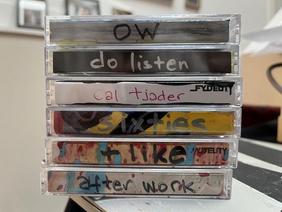 A stacked tower of six Norelco  cases, shot spine-on so you can read the labels:

"OW" — dark background, black lettering
"do listen" — dark background, white lettering
"Cal Tjader" — on a white FYDELITY-branded insert, magenta lettering
"sixties" — bold yellow lettering over a black/purple/red painted spine
"+ like" — black lettering over a cream spine splattered with blue and red paint, also FYDELITY-branded
"after work" — black lettering over a similar cream/blue/red splatter spine

The spla…