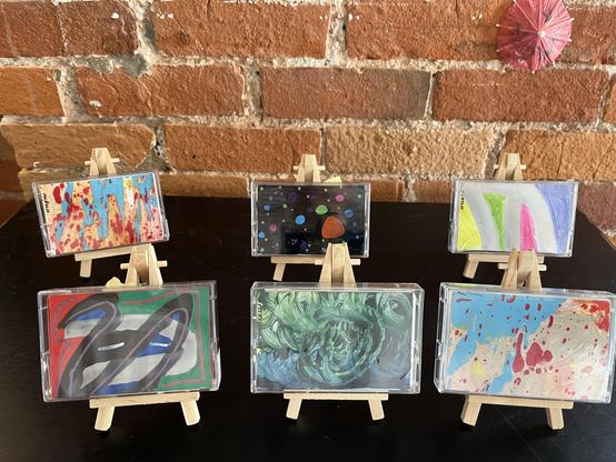 Six Norelco cases displayed face-out on mini easels against a brick wall:

Top left: cream/tan case with red splatter and blue drips (matches the "after work" splatter style)
Top center: black background with a scattering of colorful painted dots/circles — almost a night-sky or planetary look
Top right: white case with bold green, pink, and lavender curved bands, plus yellow at the edge
Bottom left: green background with a bold abstract design — black/blue swirl shapes outlined in red, with a r…