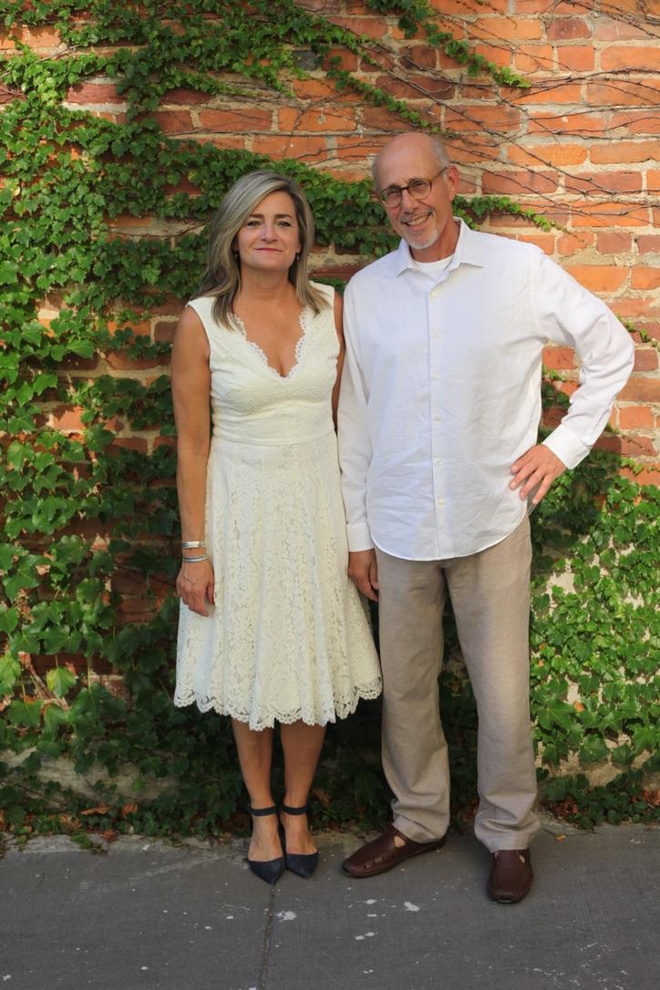 Photo of me and Lynn standing close together in the alley behind our loft, arms linked, smiling at the camera, in front of a red brick wall covered in climbing ivy with loose diagonal vine trails and some dry, leafless tendrils woven through the green.

On the left, Lynn, with shoulder-length blonde-streaked hair, wears a sleeveless cream lace dress with a scalloped V-neckline, fitted bodice, and a full knee-length skirt with a scalloped hem. What a beautiful bride! She accessorizes with a couple of thin bracelets and small earrings, and wears dark navy ankle-strap heels.

On the right, itsa me!, a man with a balding head, glasses, and a neatly trimmed white beard wearing a long-sleeved white button-down shirt with the cuffs rolled partway up, tan linen trousers, and brown leather sandals. I'm standing with one hand on my hip, smiling broadly.

We're standing on a plain concrete sidewalk in soft, warm natural light, suggesting late afternoon or early evening. The mood is casual and celebratory, a candid portrait taken just after a wedding ceremony.

ALT mostly by Claude