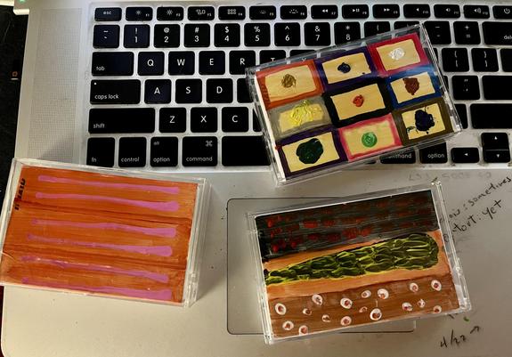Three hand-painted cassette tapes are arranged on top of a laptop keyboard and trackpad. The top tape features a nine-square grid pattern, each square a different color combination (rust/tan, purple/tan, red/tan on the top row; gray-green/tan, black/tan, blue/brown on the middle; purple/dark green, pink/green, olive/navy on the bottom), with a small raised blob of paint in a contrasting color dotted in the center of each square — giving it a tactile, almost braille-like quality. The bottom left…
