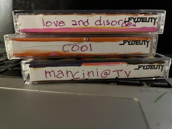 The same three cassettes are now stacked and shown from the spine/label side, resting against the laptop's keyboard near the command and option keys. Each spine is a FYDELITY-brand cassette label, hand-painted along the edges with the same colors as their respective covers, and titled in purple marker cursive/print handwriting. From top to bottom: "love and disorder" (pink and mustard painted edge), "cool" (orange painted edge, with a small drawn squiggle/line under the text), and "mancini @ TV…