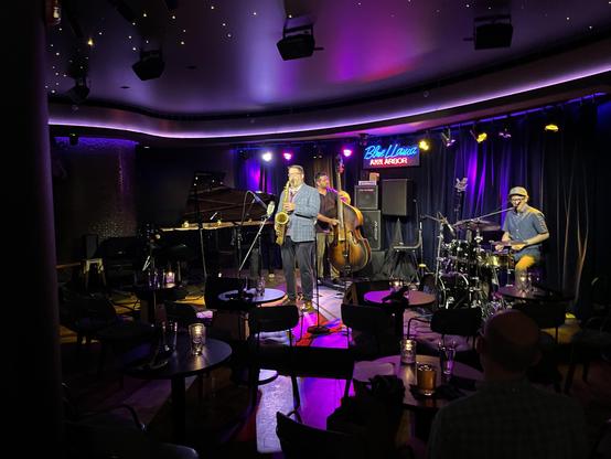 A photo of a trio mid-set at the Blue Llama. Saxophonist Andrew Bishop stands front and center under the spotlight, mid-phrase, with bassist Andy Peck just behind him working a rich, warm tone on the upright, and drummer Jess Kramer off to the right, brushes in hand, tucked behind a compact kit. The club's signature neon "Blue Llama Ann Arbor" sign glows blue and red on the back wall, and a grand piano sits unattended stage left — a trio format, just like Rollins' classic Vanguard session with …