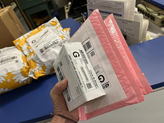 A hand holds up a small stack of USPS-labeled packages at a post office counter. The front-most package is a small package with a "USPS Ground Advantage" Click-N-Ship label, mailed from 48104. Behind it are a couple of pink bubble mailers, also with USPS labels. In the background sit a few more packages waiting to be shipped — including a yellow floral-print mailer with a daisy pattern — on a blue counter, along with some paperwork, a power strip, and other office equipment.

ALT by Claude