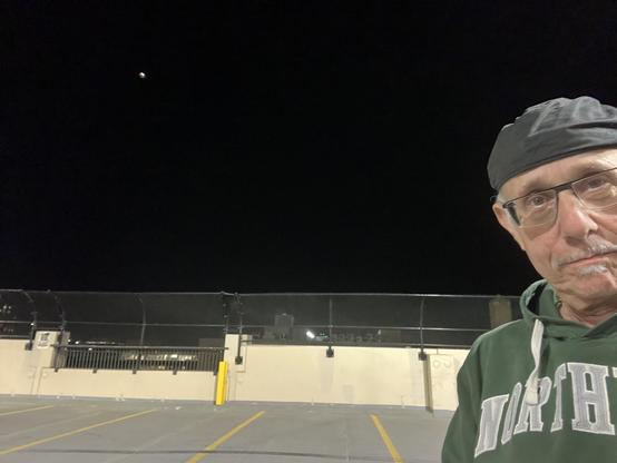 It's a nighttime selfie from the top deck of the parking structure — empty rows with yellow lane markings, a chain-link and rail barrier along the edge, and the flat rooftops and scattered lights of Ann Arbor beyond. I’m on the right in a dark cap, glasses, and a green Northern hoodie, looking straight into the camera. Up in the black sky to the upper left is the partially eclipsed moon, showing that faint reddish-copper "blood moon" tint against the darkness — a nice small but clear detail aga…