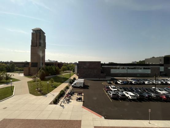 The view from my office window.

A wide daytime view over U-M's North Campus, looking out from a few floors up. On the left stands Baits Tower (the brick clock/bell tower with its distinctive stepped, angular design), rising above the surrounding plaza and lawns. Below and around it, brick-paved walkways and green lawns with young trees lead across the diag toward a low brick building in the middle distance, part of the Duderstadt Center, with a glass-walled corridor section.

In the foreground…
