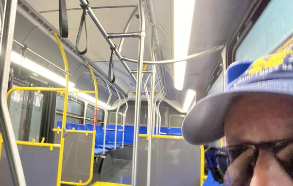 Riding the Northwood bus toward North Campus, snapping a selfie mid-ride. The photo's taken from a low angle looking toward the back of the bus, with the interior's yellow stanchions and blue seats filling most of the frame, and the ceiling grab handles dangling above. Someone in a cap and sunglasses is visible close to the camera on the right, and there's a bit of motion blur from the bus moving - the whole thing has that classic "quick pic on public transit" feel, empty seats and all.

ALT by…