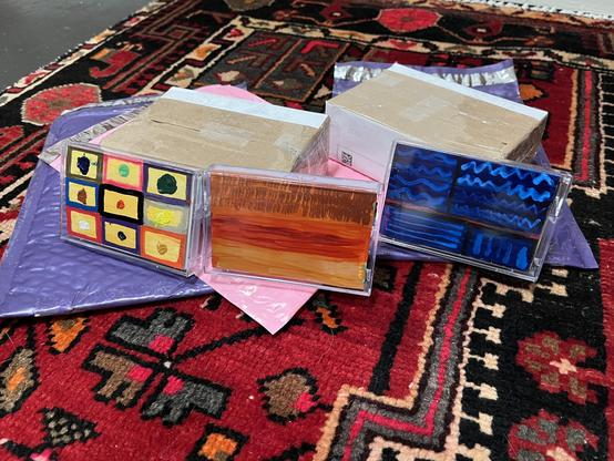 Three cassette tapes lean upright against each other in the foreground, each hand-painted with distinct designs. The left one has a nine-square grid pattern in mustard, pink, purple, and blue frames, each holding a different colored paint blob. The middle tape has a warm, streaky landscape in oranges, rust, and browns — almost like a desert sunset — with "OW" written near the top. The right tape is painted in deep blues with wavy and zigzag lines across four panels, giving it a night-sky or oce…