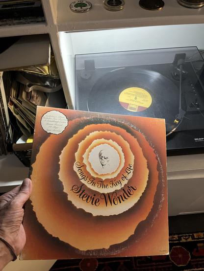 Songs in the Key of Life came out September 28, 1976, exactly 50 years ago. I’m holding up the gatefold sleeve, with its warm orange-and-amber sunburst of concentric, ragged-edged rings around a small pen-and-ink portrait at the center. The title curves along the inner ring and Stevie Wonder's name in flowing script below it. A round sticker in the upper left reads "Collector's Album, Includes Two Records, A Something's Extra Bonus Record, 24-Page Lyric Booklet." That's the original double LP p…