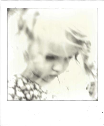 Extreme close-up Polaroid of a child's face, heavily blurred and overexposed so the image looks dreamlike. The child looks downward, with a dark smudge where the eyes are and the nose and mouth softly visible. Wisps of blond hair fall in dark, feathery streaks across the top and right of the frame. At lower left, a geometric diamond-lattice pattern on the child's clothing is the sharpest detail in the picture. Most of the frame fades to bright, creamy white, with faint rainbow-like chemical swi…