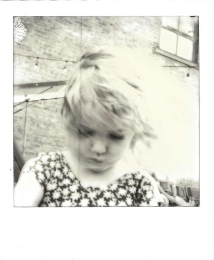 Black-and-white Polaroid of a young child with short, tousled blond hair, looking down with eyes nearly closed. The image is soft and blurred, as if the child moved during the exposure. The child wears a sleeveless dress with a bold floral pattern in dark and white. Behind them is a weathered brick wall with a string of café lights sagging across it and a multi-paned window at upper right. The right side of the frame is washed out to a pale cream glow, and small swirled chemical artifacts mark …
