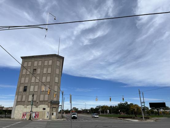 This is a wide-angle, upward-looking shot dominated by sky. The top two-thirds is a sweep of wispy cirrus cloud that ends in a crisp, diagonal edge, with clean deep blue below it. A heavy black utility cable cuts across the top of the frame, along with a mast arm holding a hanging streetlight.

On the left is the C·P·A Building, a narrow five-story masonry building with a dentiled cornice, rows of small rosette ornaments, and a thin antenna on the roof. Almost every window is covered in weather…