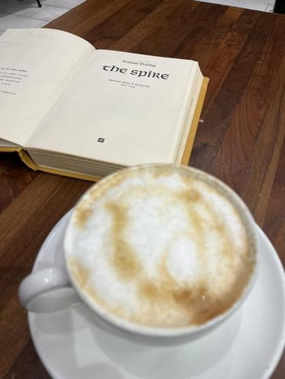 A hardcover copy of William Golding’s The Spire lies open to its title page (Harcourt, Brace & World, New York) on a dark wood table. In the foreground, slightly out of focus, is a white cup of cappuccino on a saucer, with a swirl of milk foam.
ALT by Claude