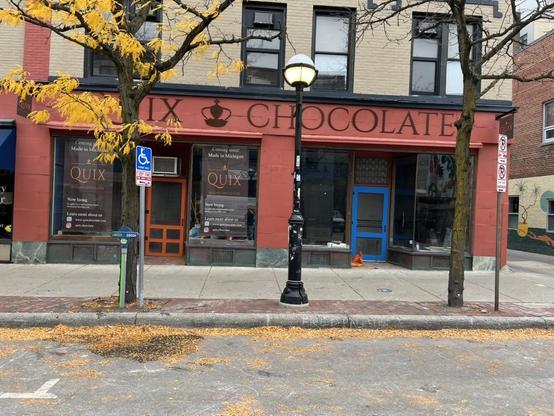 Street-level photo of a terracotta-red brick storefront with a large brown sign reading “QUIX CHOCOLATE,” a small crown emblem between the two words. A tree with bright yellow autumn leaves stands at the left, partly covering the sign, and a black globe street lamp stands at center. Two large display windows hold dark brown posters with the word “QUIX” in an elegant serif typeface, “Belgian Chocolates” beneath it, and the text “Coming soon! Made in Michigan,” plus “Now hiring” with a website an…