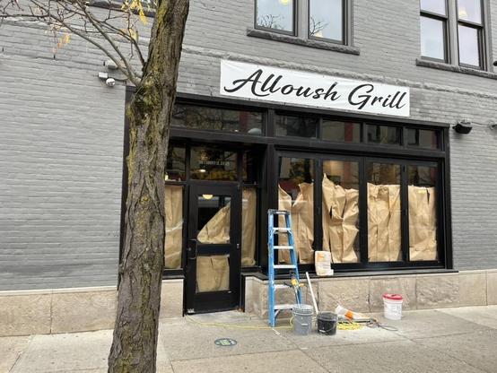Photo of the street-level facade of a building with gray-painted brick and a stone base. A white banner above the entrance reads “Alloush Grill” in black cursive script. Below it, black-framed windows and a glass door are covered on the inside with brown kraft paper, hiding the interior, a sign that the space is being prepared for opening. Small gold lettering on the door glass reads “106 E Liberty St, Ste 120.” In front, a blue stepladder leans against the stone base. On the sidewalk sit two g…