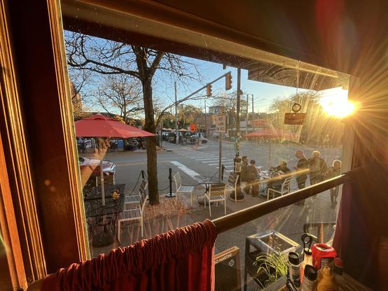Late-afternoon sun is pouring through the window, with a starburst flare just above the right edge of the frame and a golden glow washing over the glass. The window itself is a little smudged and spotted, which catches the light and adds to the warm haze.

Outside is the corner of Liberty and Ashley, with a "No Turn on Red" sign and a "First St Detour" sign in orange. Traffic lights hang from a mast arm over the crosswalk, and a bare-branched tree with a few yellow leaves stands in the foregrou…