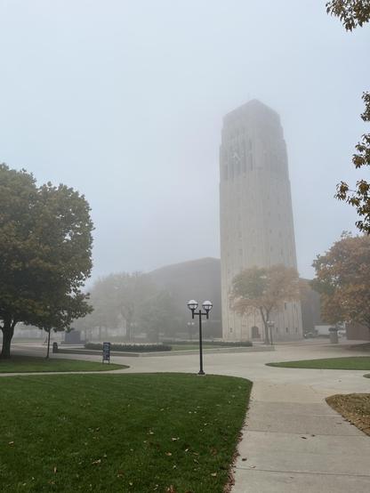 Burton Tower is wrapped in thick fog, its upper third dissolving into a pale gray-blue sky so the top seems to disappear. The limestone shaft rises in tall vertical bands with stepped, Art Deco setbacks. Narrow windows stack up its face, and a clock face is faintly visible on the upper section, with the louvered bell-chamber openings above it barely discernible through the haze.

The foreground is quiet and muted. A curving concrete walkway sweeps from the lower right toward the tower, bordered…